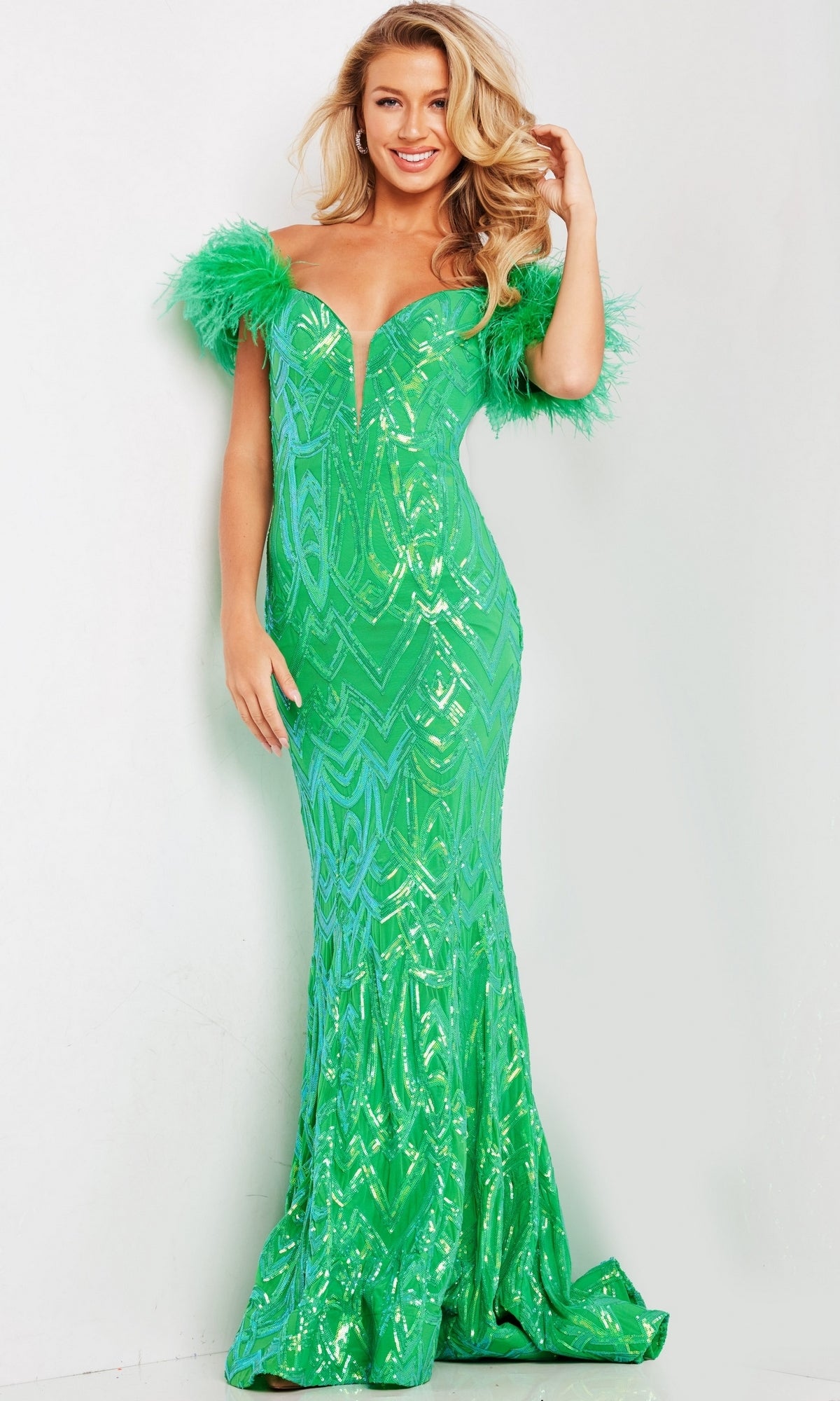A woman stands and smiles against a plain background, wearing the Jovani Feather-Trim Long Sequin Prom Dress 23383—an elegant green, off-the-shoulder floor-length gown with feathered trim, shimmering geometric pattern, and fitted silhouette.